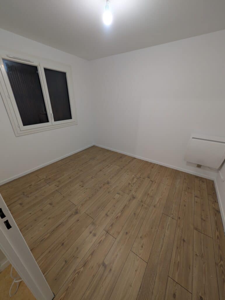 Pièce vide et lumineuse en rénovation. Murs blancs, sol en parquet clair type pin, et grande fenêtre. Présence d'un radiateur mural.